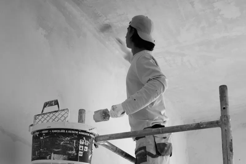 Peintre en bâtiment debout sur un échafaudage, pinceau à la main contre un mur, en noir et blanc